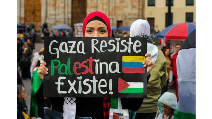 Mujer sosteniendo un cartel en una marcha que dice Gaza Resiste Palestina Existe junto a las banderas de Colombia y Palestina